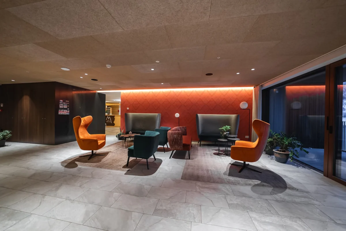 Modern hotel lobby seating area with orange and teal chairs around small tables against a red wall. Large windows to the right and plants nearby.