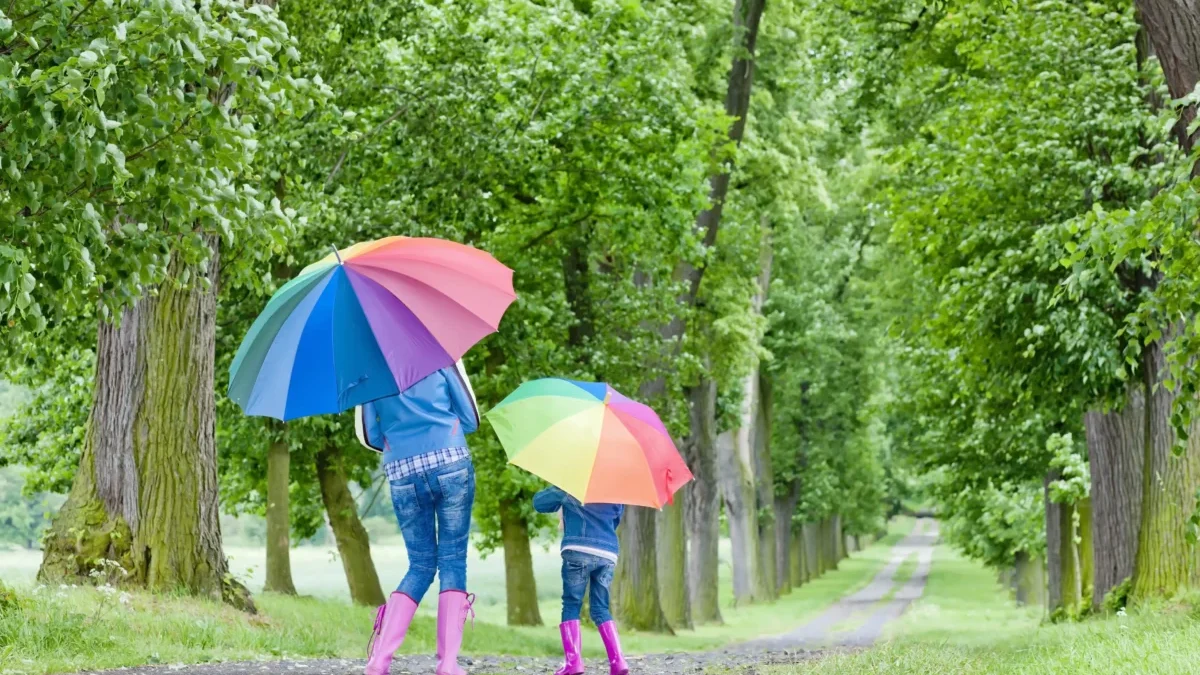 Žena s dcerou mají nad hlavami deštníky a procházejí se přírodou.