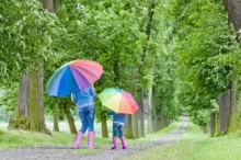Žena s dcerou mají nad hlavami deštníky a procházejí se přírodou.