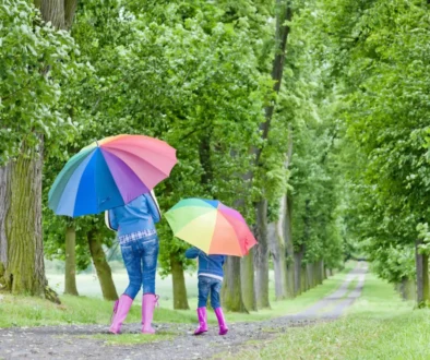 Žena s dcerou mají nad hlavami deštníky a procházejí se přírodou.