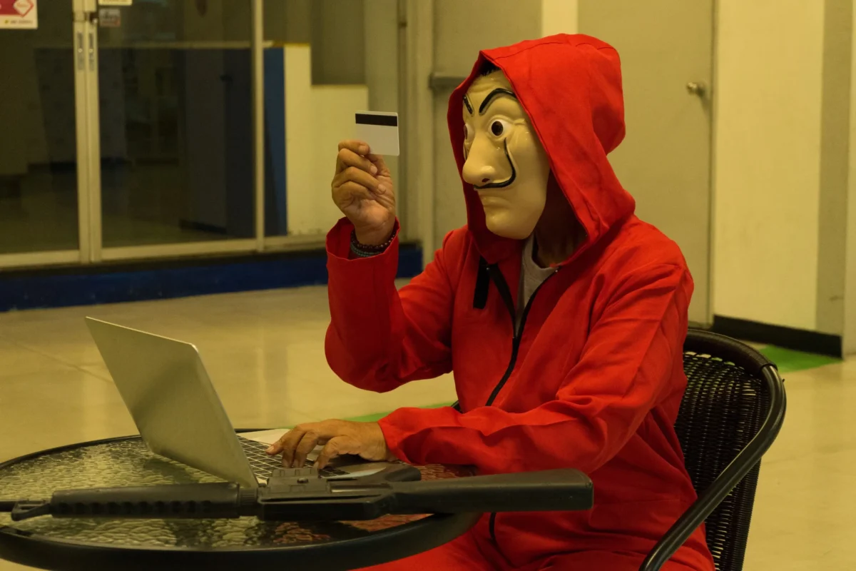 Person in a red hoodie wearing a mask sits at a table with a laptop, holding up a small card with a black stripe toward the camera.
