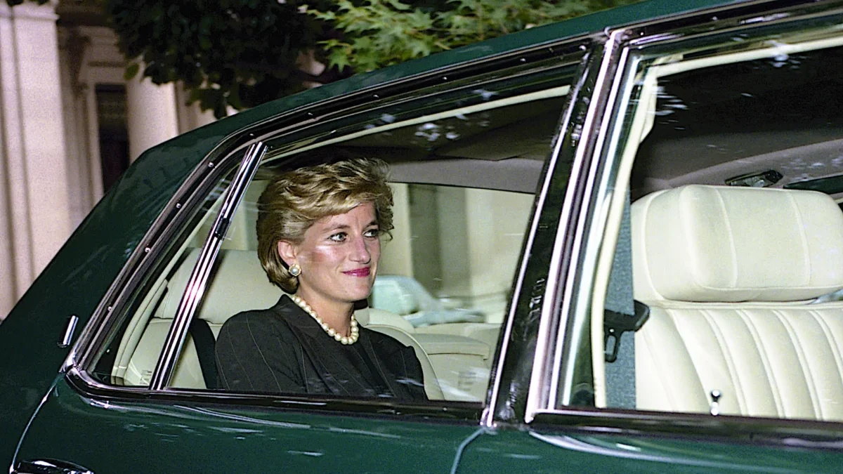 Woman with short blonde hair and pearl necklace sits in the passenger seat of a dark green car with beige interior and window down.