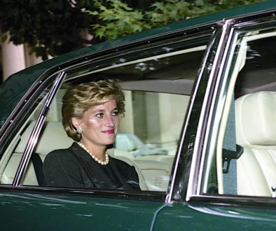 Woman with short blonde hair and pearl necklace sits in the passenger seat of a dark green car with beige interior and window down.