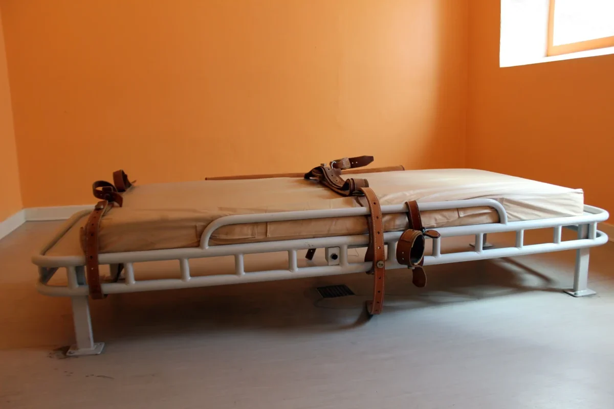 A hospital gurney with a beige mattress and brown straps sits in an empty orange-walled room by a window.