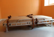 A hospital gurney with a beige mattress and brown straps sits in an empty orange-walled room by a window.