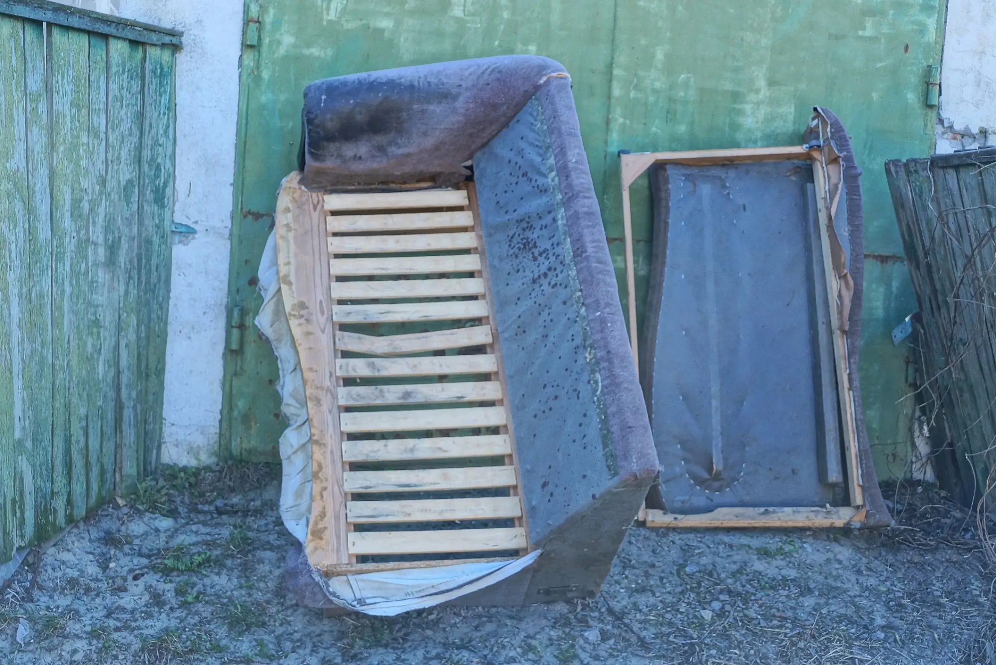 Discarded mattresses and bed frames stacked against a weathered green fence in an outdoor area.”] ,