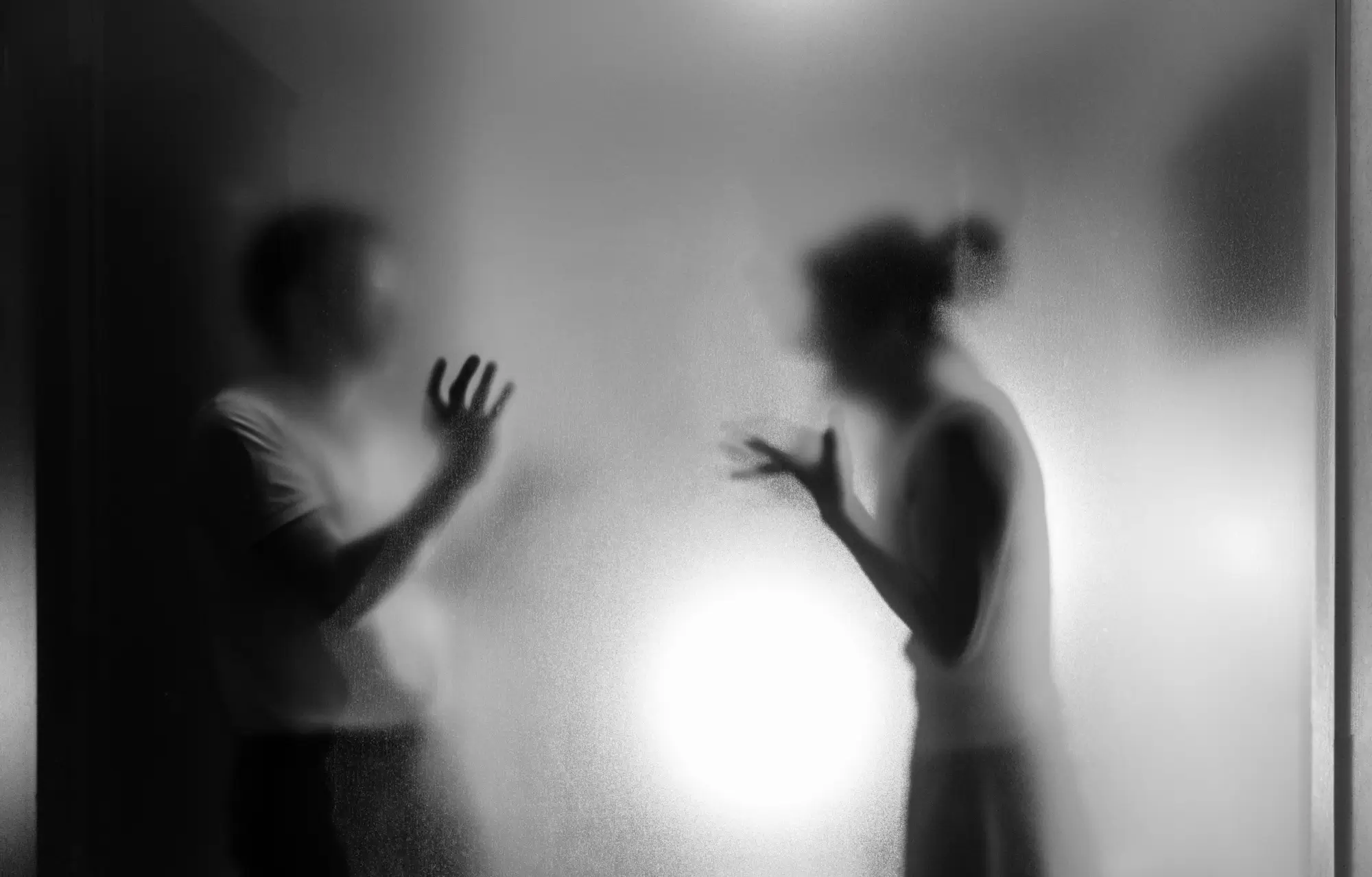 Two blurred silhouettes press their hands and faces against a frosted glass panel, backlit by a bright light behind them.