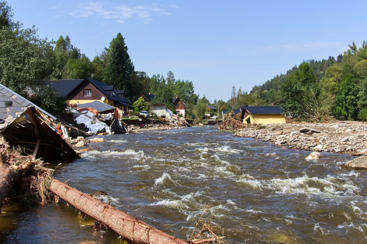Rushing river beside a damaged suburban street, with collapsed houses and debris along the bank and trees in the background.