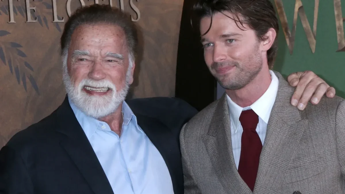 Two men pose for a photo at an event; the older man with a white beard smiles beside a younger man in a brown checkered suit and red tie, both in formal attire.