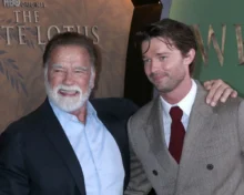 Two men pose for a photo at an event; the older man with a white beard smiles beside a younger man in a brown checkered suit and red tie, both in formal attire.