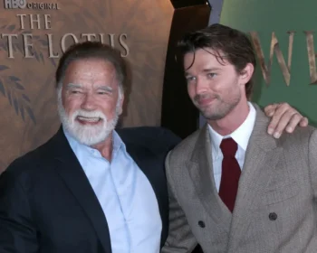 Two men pose for a photo at an event; the older man with a white beard smiles beside a younger man in a brown checkered suit and red tie, both in formal attire.
