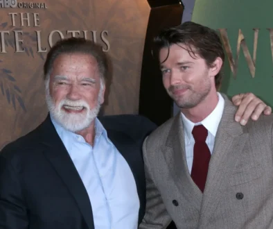 Two men pose for a photo at an event; the older man with a white beard smiles beside a younger man in a brown checkered suit and red tie, both in formal attire.