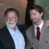 Two men pose for a photo at an event; the older man with a white beard smiles beside a younger man in a brown checkered suit and red tie, both in formal attire.