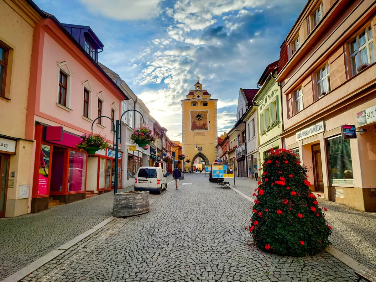 Colorful European street with pastel buildings, cobblestone pavement, and a yellow clock tower in the distance; planters with red flowers line the sidewalks.