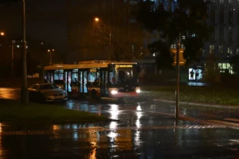 Nighttime street scene with a bus stopped at a curb in rainy weather; headlights reflect on wet pavement and streetlights glow in the scene.