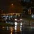 Nighttime street scene with a bus stopped at a curb in rainy weather; headlights reflect on wet pavement and streetlights glow in the scene.