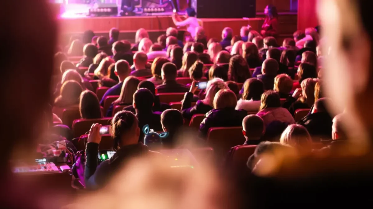 Audience seated in a dim theater watching a stage performance with pink-purple lighting, viewed from the back of the room.