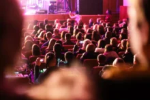 Audience seated in a dim theater watching a stage performance with pink-purple lighting, viewed from the back of the room.