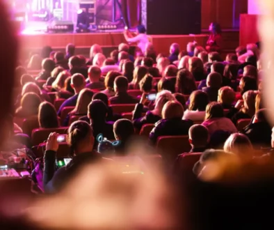 Audience seated in a dim theater watching a stage performance with pink-purple lighting, viewed from the back of the room.