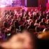 Audience seated in a dim theater watching a stage performance with pink-purple lighting, viewed from the back of the room.