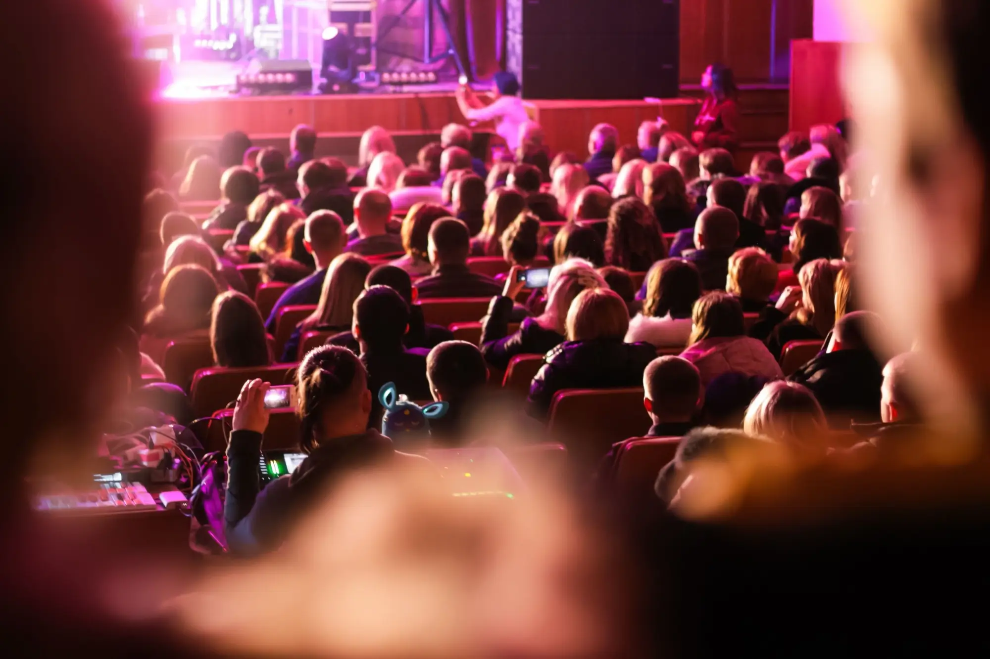 Audience seated in a dim theater watching a stage performance with pink-purple lighting, viewed from the back of the room.