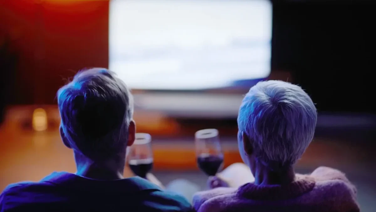 Two people with gray hair sit on a couch facing a bright screen in a dark room, wine glasses on the coffee table in front.