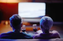 Two people with gray hair sit on a couch facing a bright screen in a dark room, wine glasses on the coffee table in front.