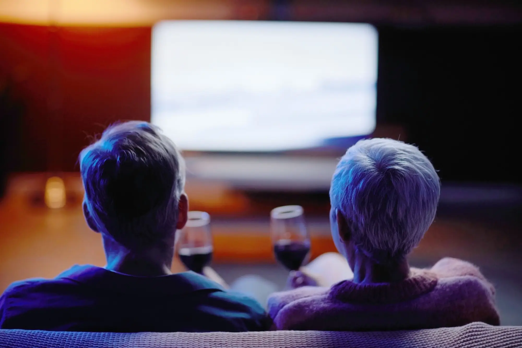 Two people with gray hair sit on a couch facing a bright screen in a dark room, wine glasses on the coffee table in front.