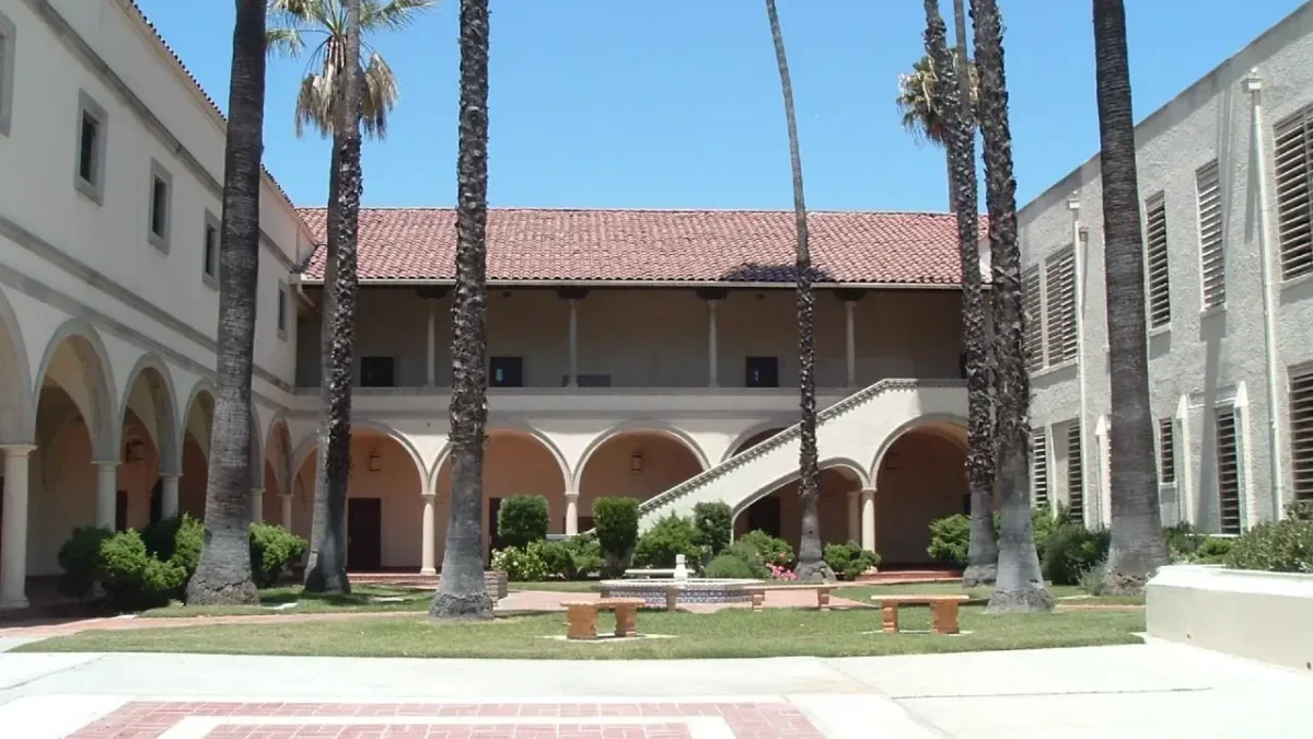 Sunny campus courtyard with tall palm trees, arched walkways, and a central fountain surrounded by benches.