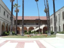 Sunny campus courtyard with tall palm trees, arched walkways, and a central fountain surrounded by benches.