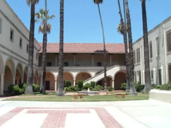 Sunny campus courtyard with tall palm trees, arched walkways, and a central fountain surrounded by benches.