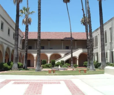 Sunny campus courtyard with tall palm trees, arched walkways, and a central fountain surrounded by benches.