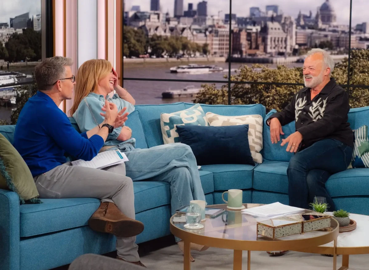 Three adults sit on a blue sectional sofa chatting and laughing in a TV talk-show set with a city skyline backdrop and a coffee table in front.