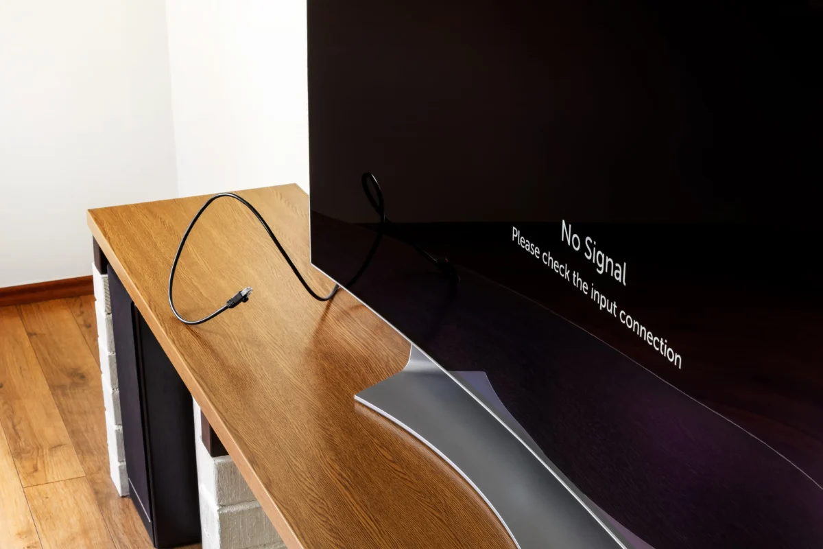 Flat-screen TV on a wooden stand shows a 'No Signal' message on the bottom right, with a coiled HDMI cable on the desk surface.