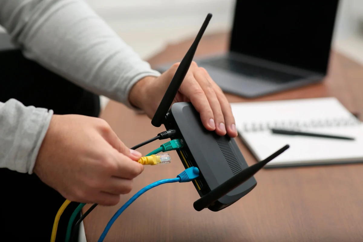 Close-up of hands connecting color-coded Ethernet cables to a networking device on a desk with a laptop in the background.
