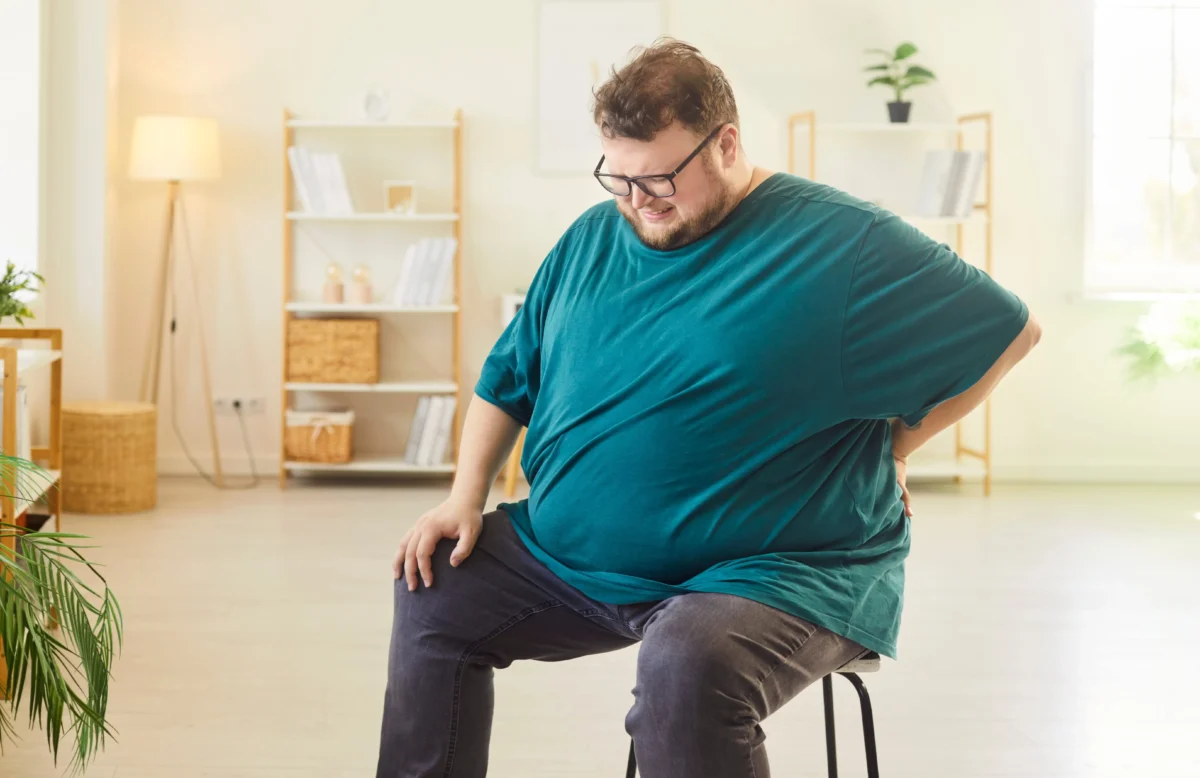 shutterstock_2702705259 1 | TVGURU.cz Man wearing a teal shirt and glasses sits on a tall stool in a bright room, looking down with a slight smile.