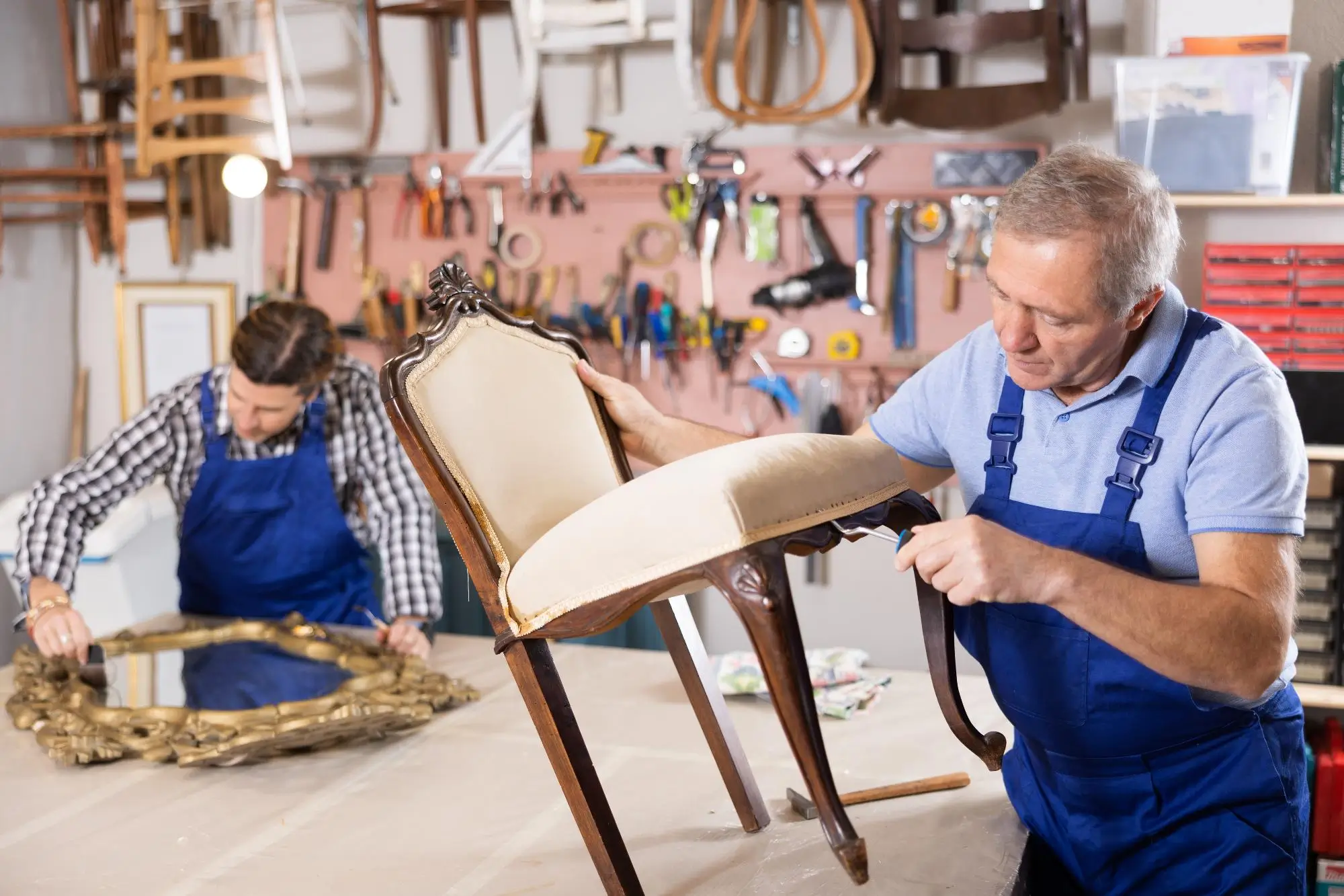Two men in blue overalls upholster chairs in a workshop filled with tools on a pegboard behind them.