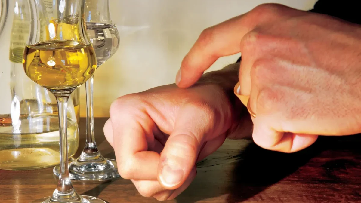 Close-up of hands wiping a wooden table with a small orange cloth beside two wine glasses and a bottle in the background.