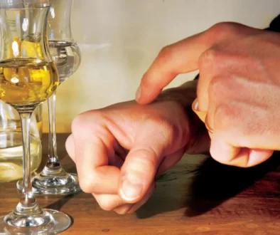 Close-up of hands wiping a wooden table with a small orange cloth beside two wine glasses and a bottle in the background.