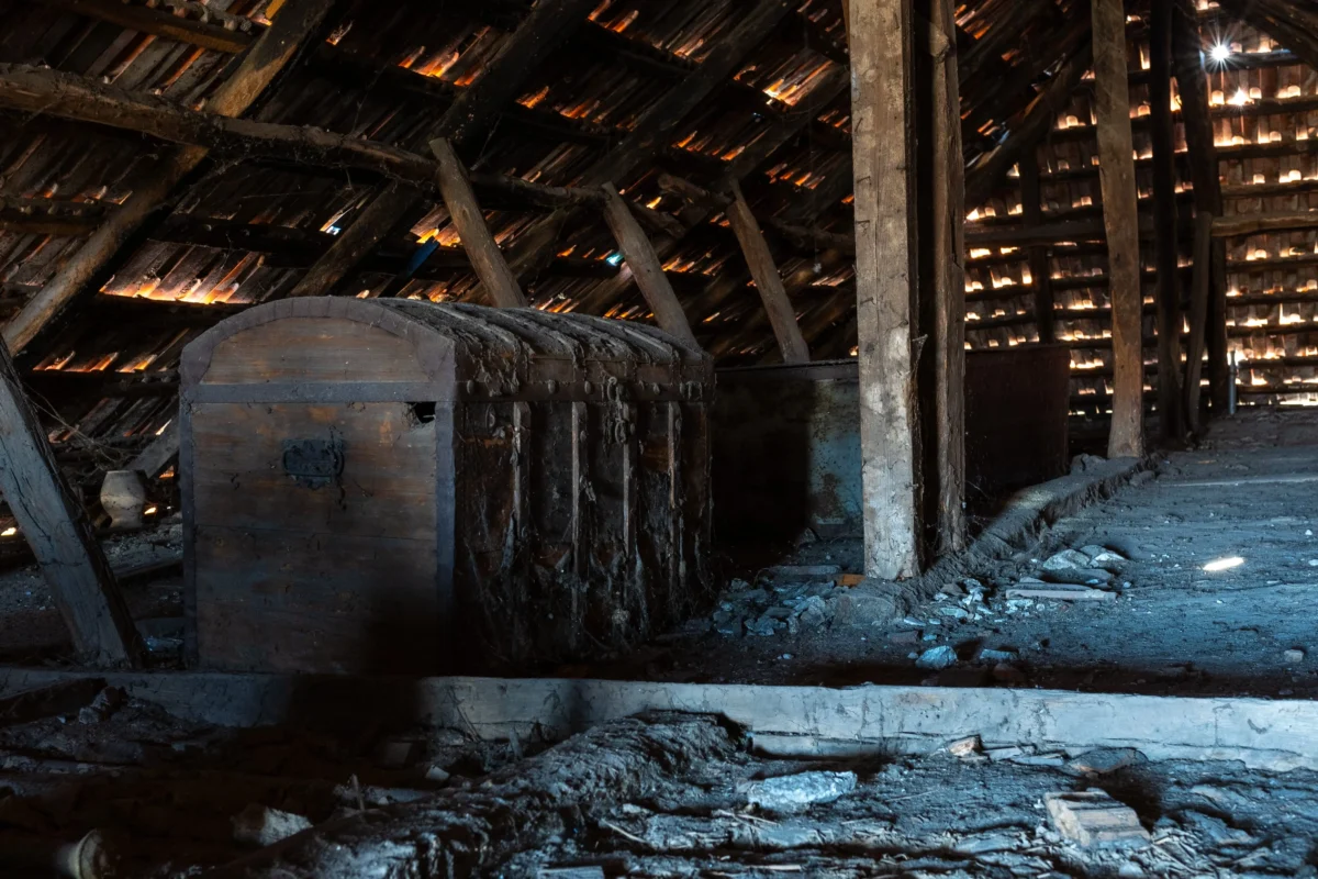 shutterstock_2422748111 1 | TVGURU.cz Dusty, abandoned attic with rough wooden beams, a large old chest, and sunlight filtering through gaps in the slatted roof.
