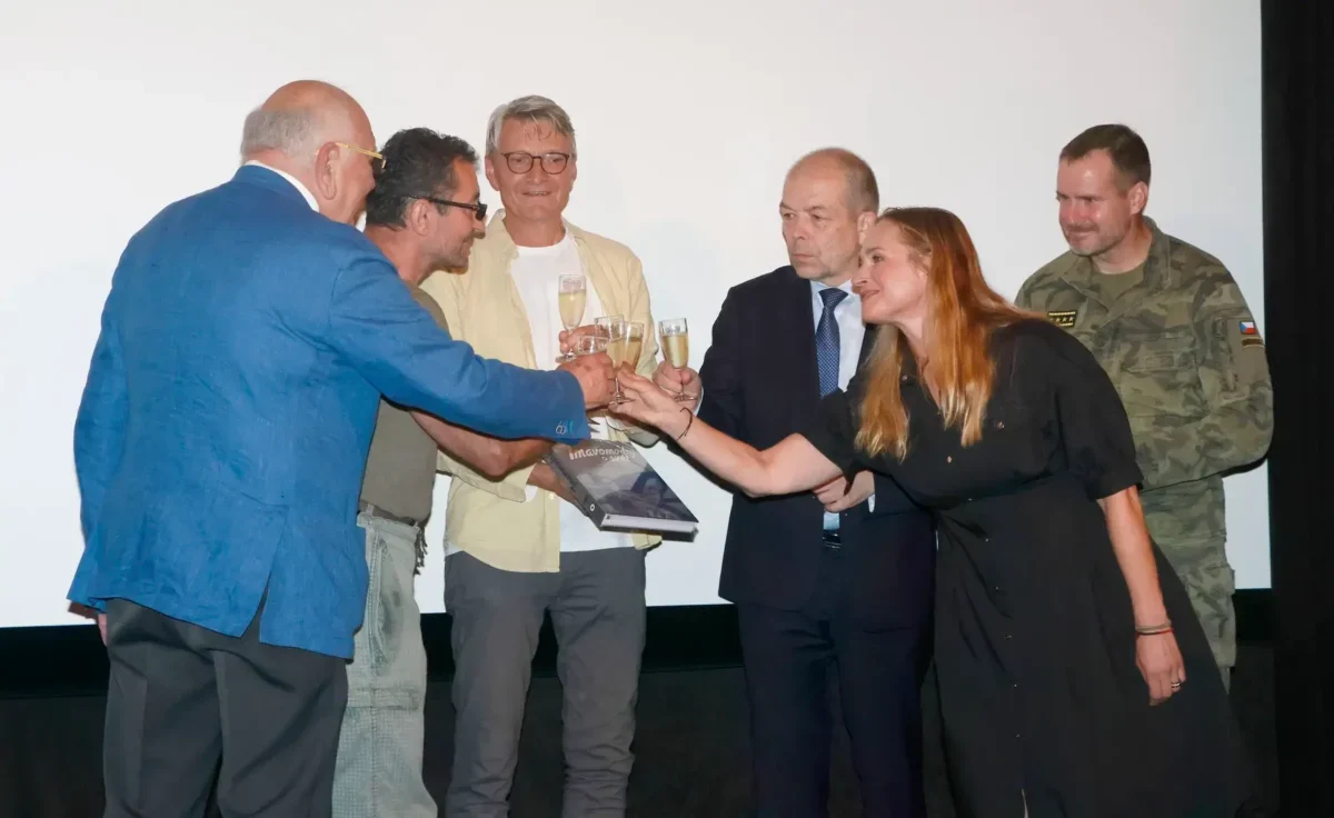 Group of six adults on a stage toasting with champagne glasses, including a man in a blue suit, a woman in black, and a soldier in camo, against a white backdrop.