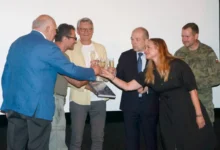 Group of six adults on a stage toasting with champagne glasses, including a man in a blue suit, a woman in black, and a soldier in camo, against a white backdrop.