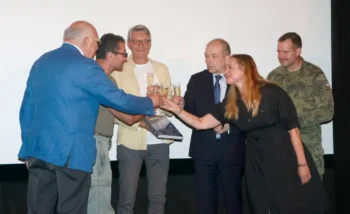 Group of six adults on a stage toasting with champagne glasses, including a man in a blue suit, a woman in black, and a soldier in camo, against a white backdrop.
