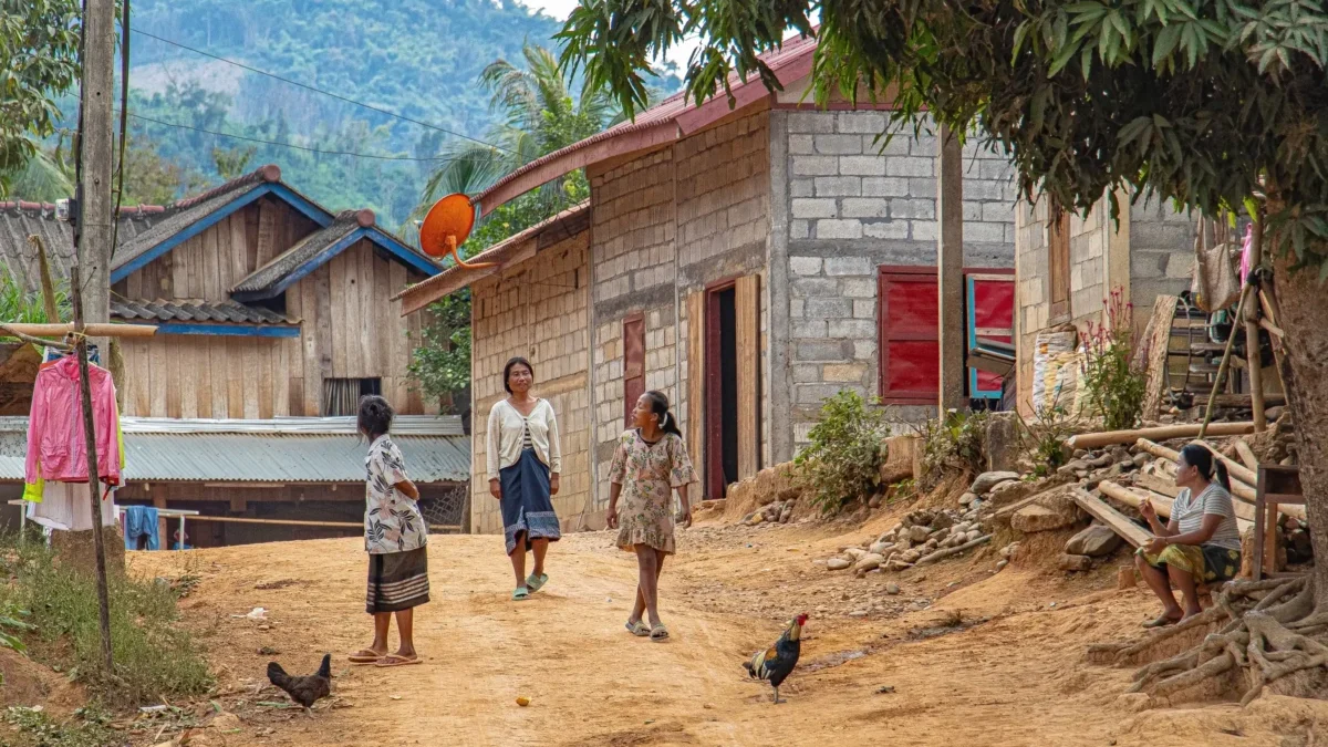 Dusty village street with women and children walking past simple brick houses; a rooster in the foreground.