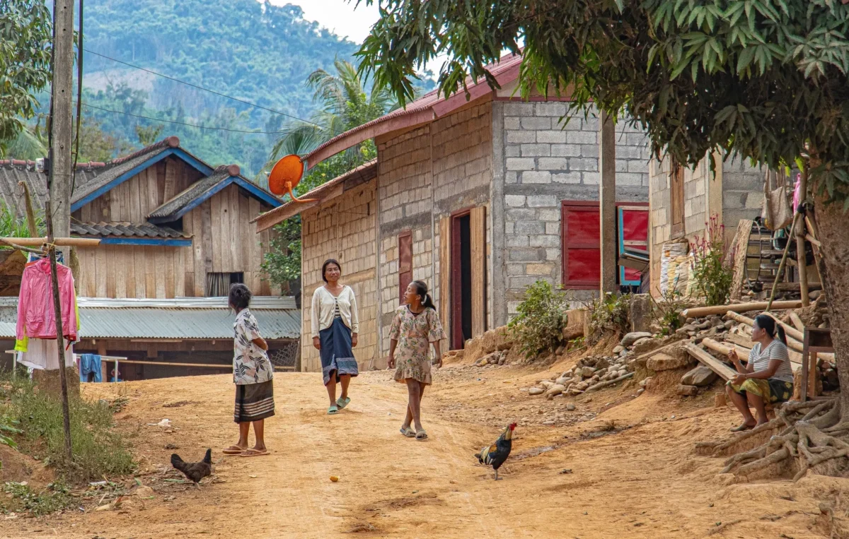 Dusty village street with women and children walking past simple brick houses; a rooster in the foreground.