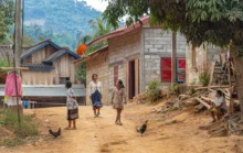 Dusty village street with women and children walking past simple brick houses; a rooster in the foreground.