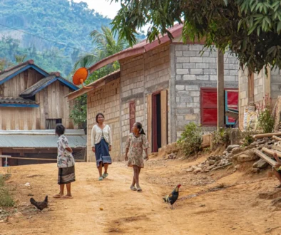 Dusty village street with women and children walking past simple brick houses; a rooster in the foreground.