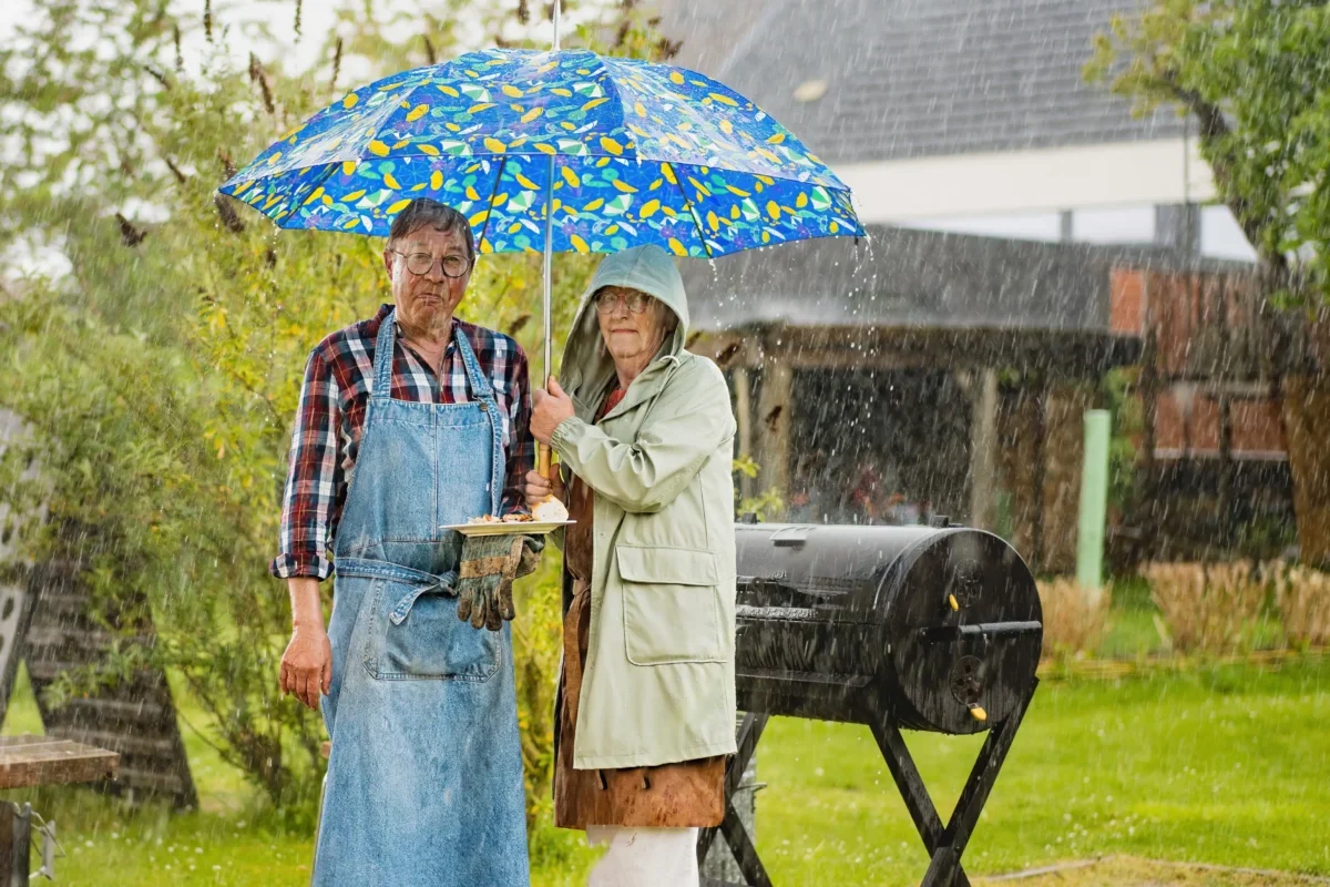 Dva lidé stojí na zahradě u grilu v dešti pod deštníkem