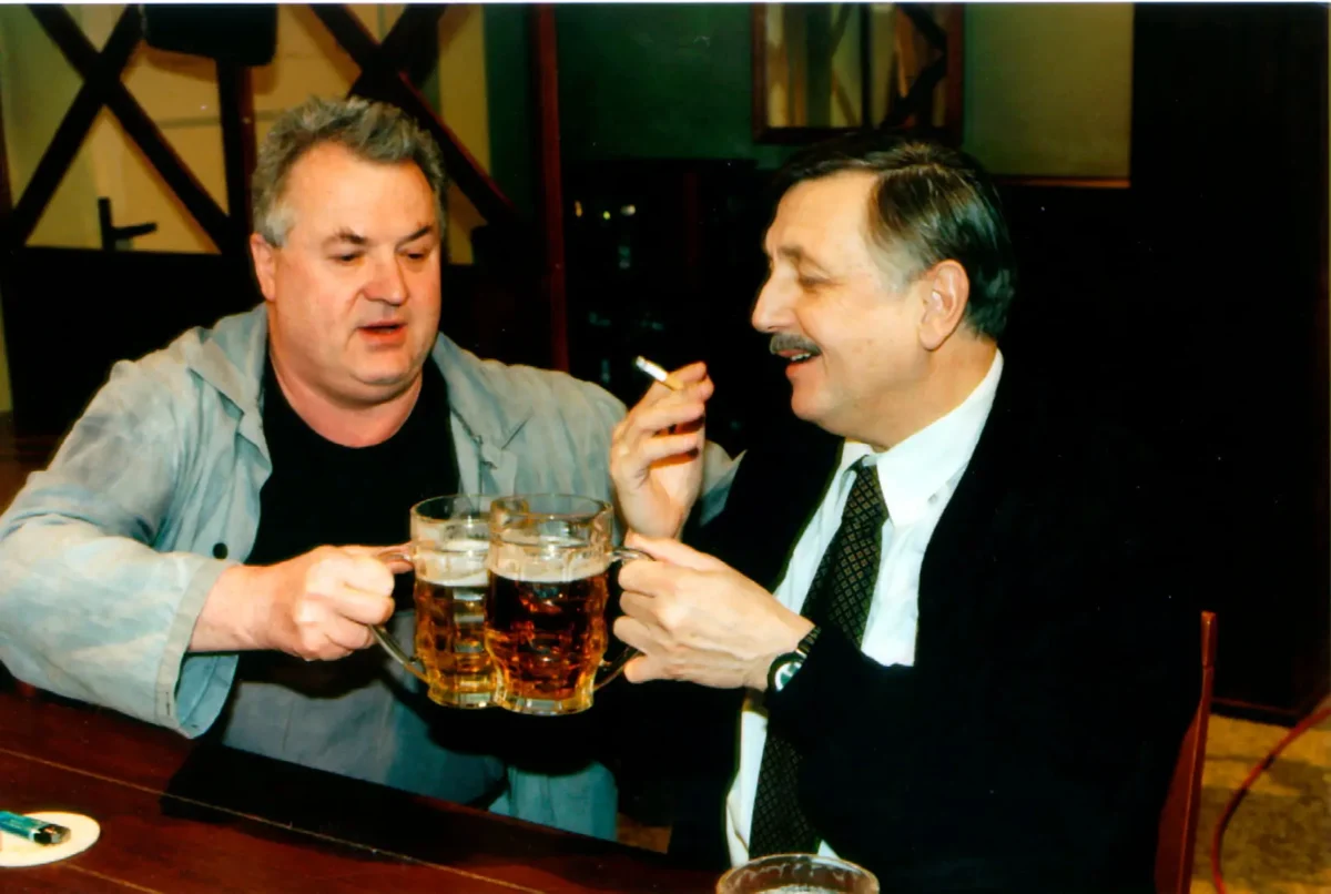 profimedia-0670681391 1 | TVGURU.cz Two middle-aged men toast with large beer mugs at a bar, one holding a cigarette and smiling at the other.