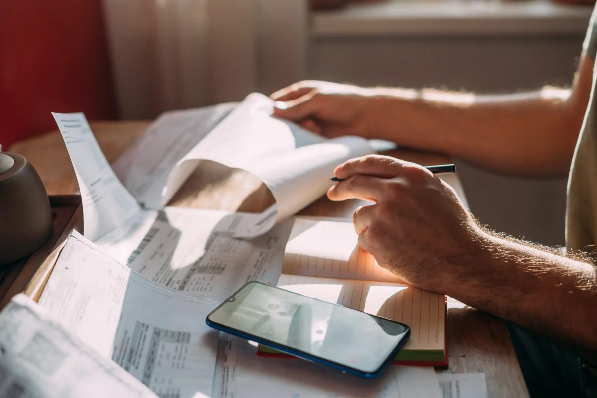 shutterstock_2505463005 1 | TVGURU.cz Hands writing on scattered receipts and documents with a pen; a smartphone rests on the desk nearby.